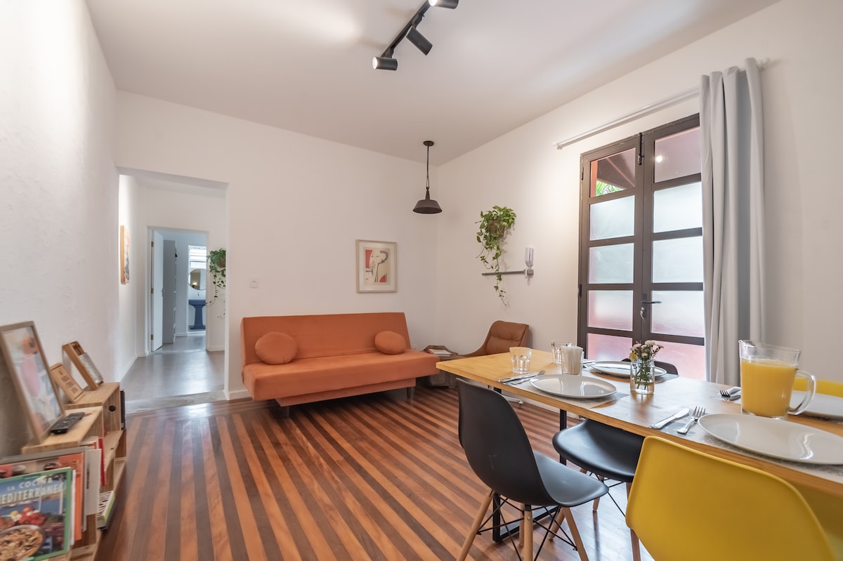 A modern dining area features a wooden table set for a meal, with chairs in various colors surrounding it. A cozy orange sofa is positioned against the wall, while sunlight streams through large glass doors, illuminating the space.
