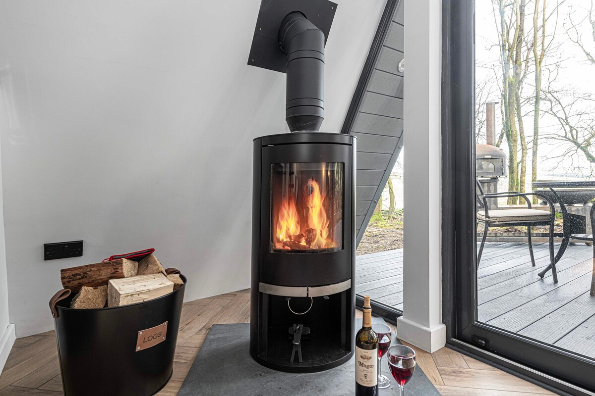 A modern log burner is situated within the cabin, featuring a glass front that displays a flickering flame. Next to it, a basket filled with logs is positioned. A small wine bottle and glass stand nearby, adding to the cozy ambiance.