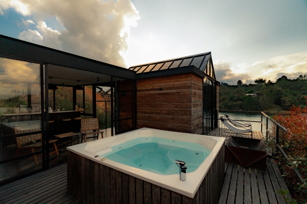 An outdoor space features a hot tub set on a wooden deck, surrounded by natural scenery. Large glass doors provide a view of the interior area, while soft clouds drift across the sky. A hammock and additional seating are visible, creating a relaxing environment.