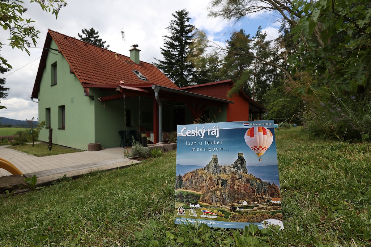 A two-story residence features a green exterior and a red-tiled roof. The well-maintained garden includes landscaped grass in the foreground. A promotional sign advertising Český ráj is prominently displayed, highlighting the area’s natural beauty and attractions.