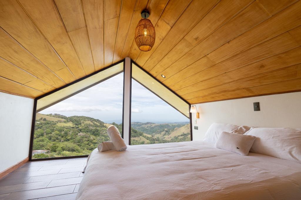 A spacious sleeping area features a large bed adorned with white bedding. A striking triangular window showcases panoramic views of rolling hills and distant mountains, while natural wood accents create a warm ambiance in the room.