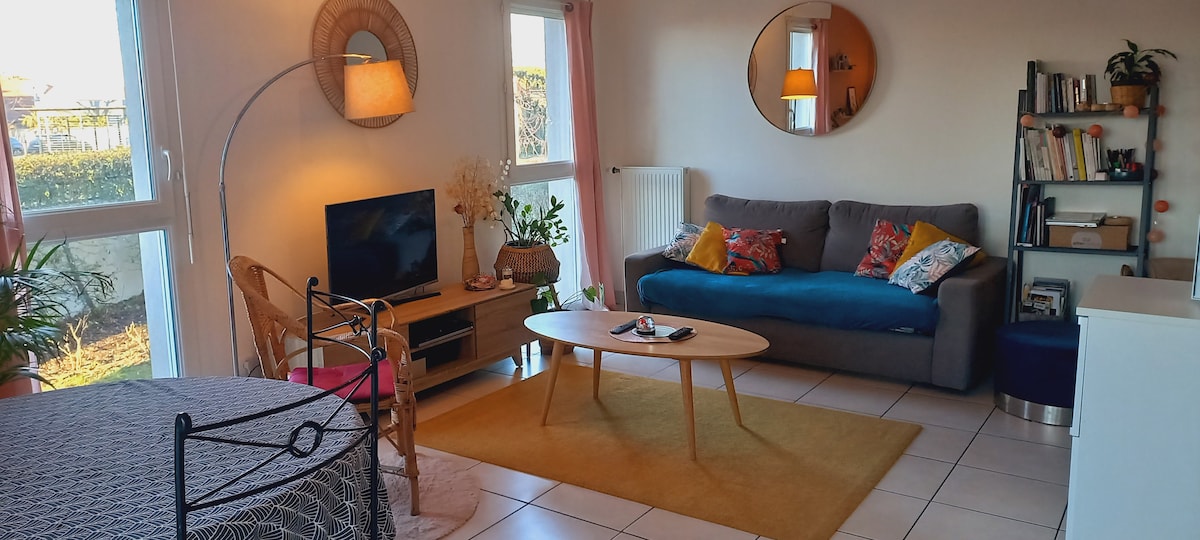 A bright living area features a comfortable sofa adorned with colorful cushions and a coffee table positioned centrally. A television is placed on a wooden console beside potted plants. A cozy seating area is highlighted by a warm rug, complemented by natural light from large windows.