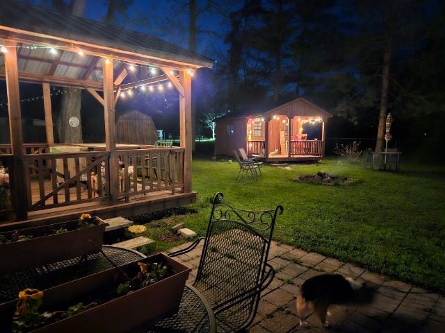 A charming outdoor area features a wooden gazebo adorned with soft lighting and seating options. A cozy cabin is visible nearby, with an inviting porch. Lush green grass surrounds a stone patio, which includes garden boxes and pathway stones leading into the tranquil setting.