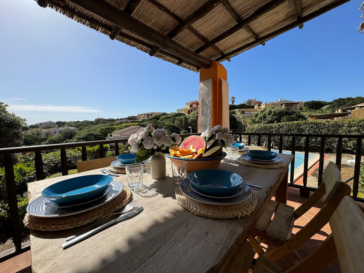 A dining table is set on a spacious veranda, featuring blue plates, woven placemats, and a floral centerpiece. The setting overlooks lush greenery and offers glimpses of nearby villas against a clear blue sky.