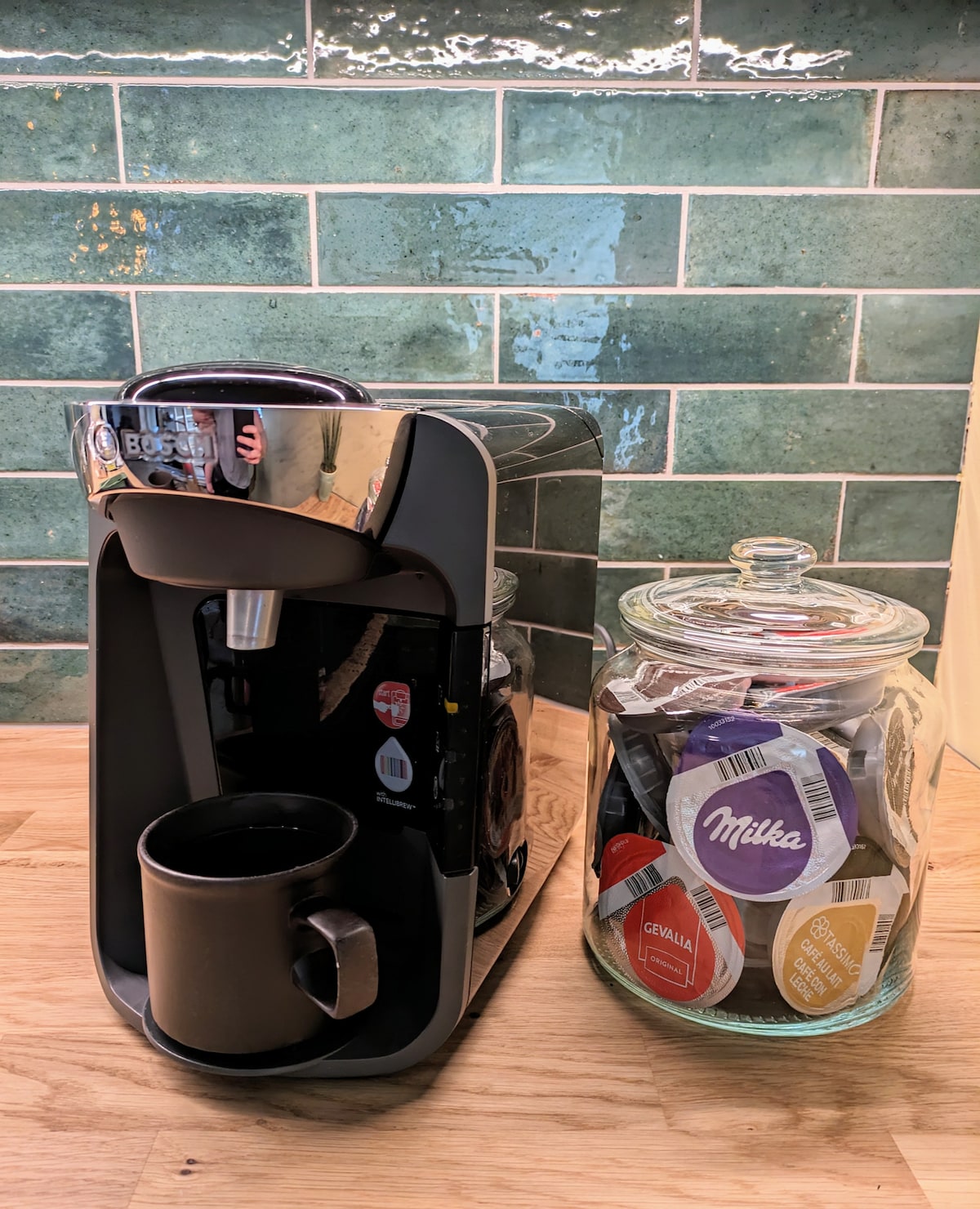 A modern coffee machine is positioned next to a glass jar filled with assorted coffee pods. A black mug is placed beneath the machine’s dispensing area, and the backdrop features a green tiled wall, enhancing the kitchen's contemporary design.