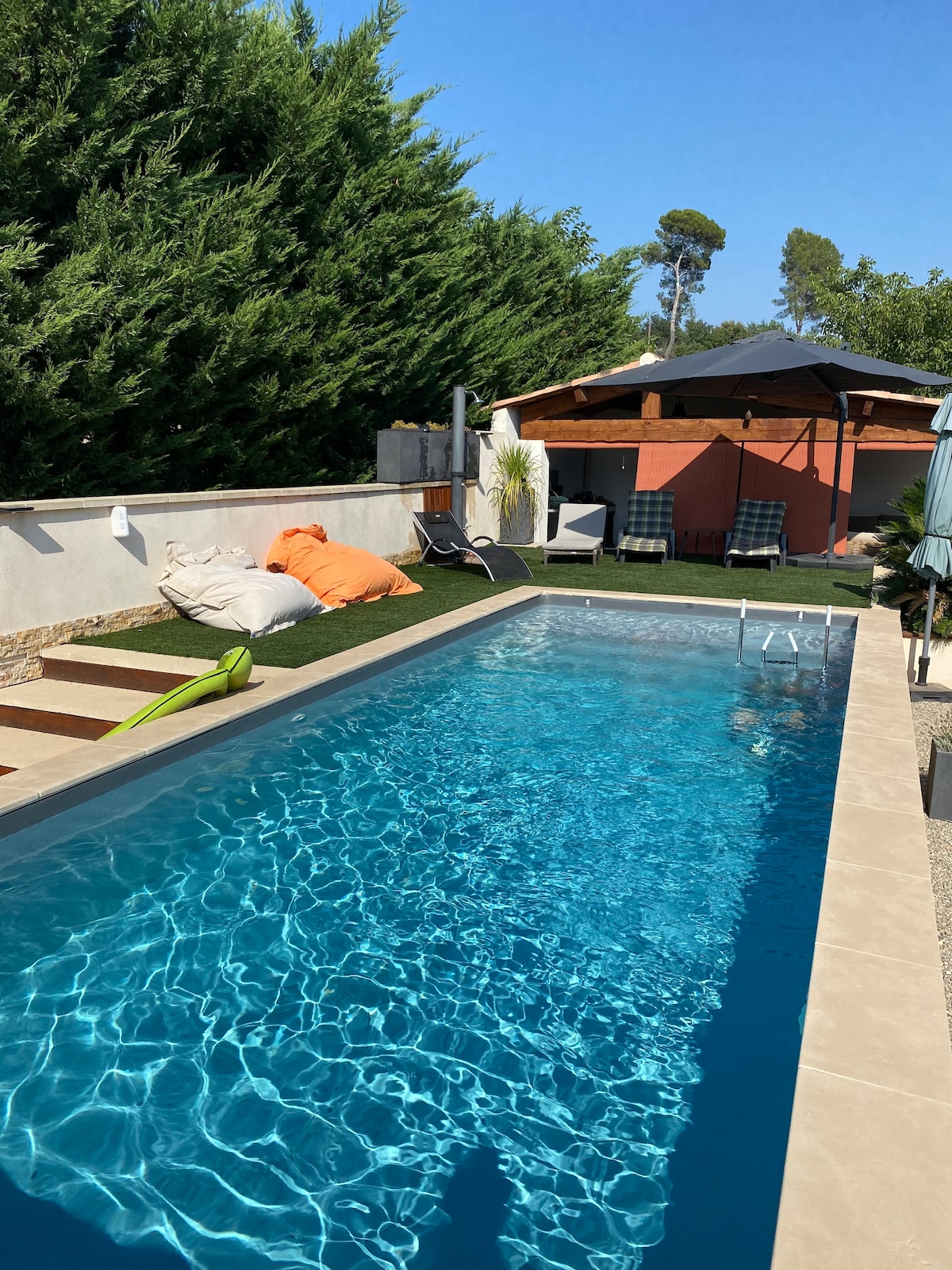 A serene swimming pool is surrounded by green hedges and features clear blue water reflecting sunlight. Comfortable lounge chairs are positioned nearby, while vibrant cushions add a splash of color. A shaded area is visible at the end of the pool, providing a shaded retreat.