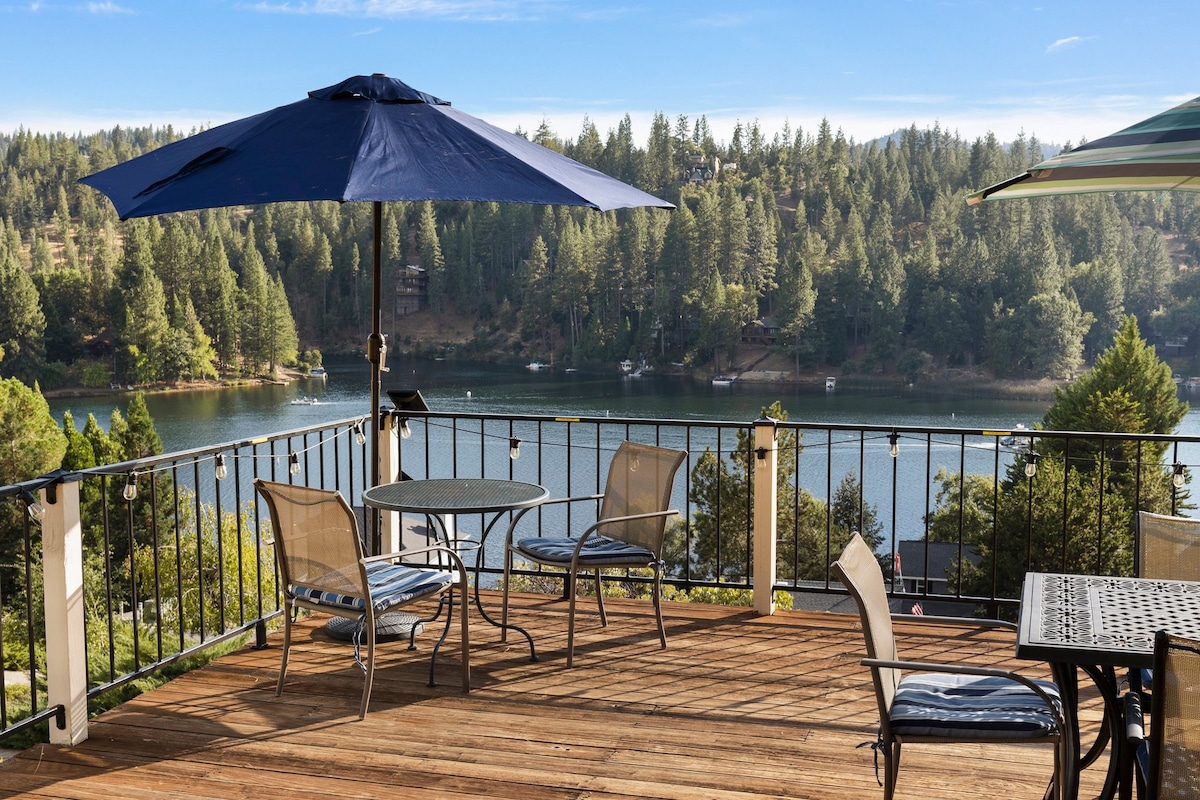 A spacious deck features a dark blue umbrella providing shade over a round table with four chairs. The wooden surface enhances the view of the lake and surrounding trees, creating a serene outdoor space ideal for relaxation.