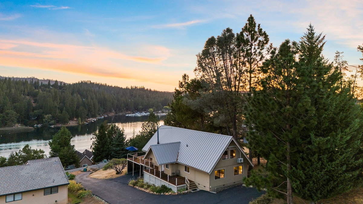 An aerial view captures a charming A-frame cabin surrounded by tall trees, with the serene Pine Mountain Lake in the background. The setting sun casts a warm glow on the landscape, highlighting the cabin's welcoming facade and spacious driveway.