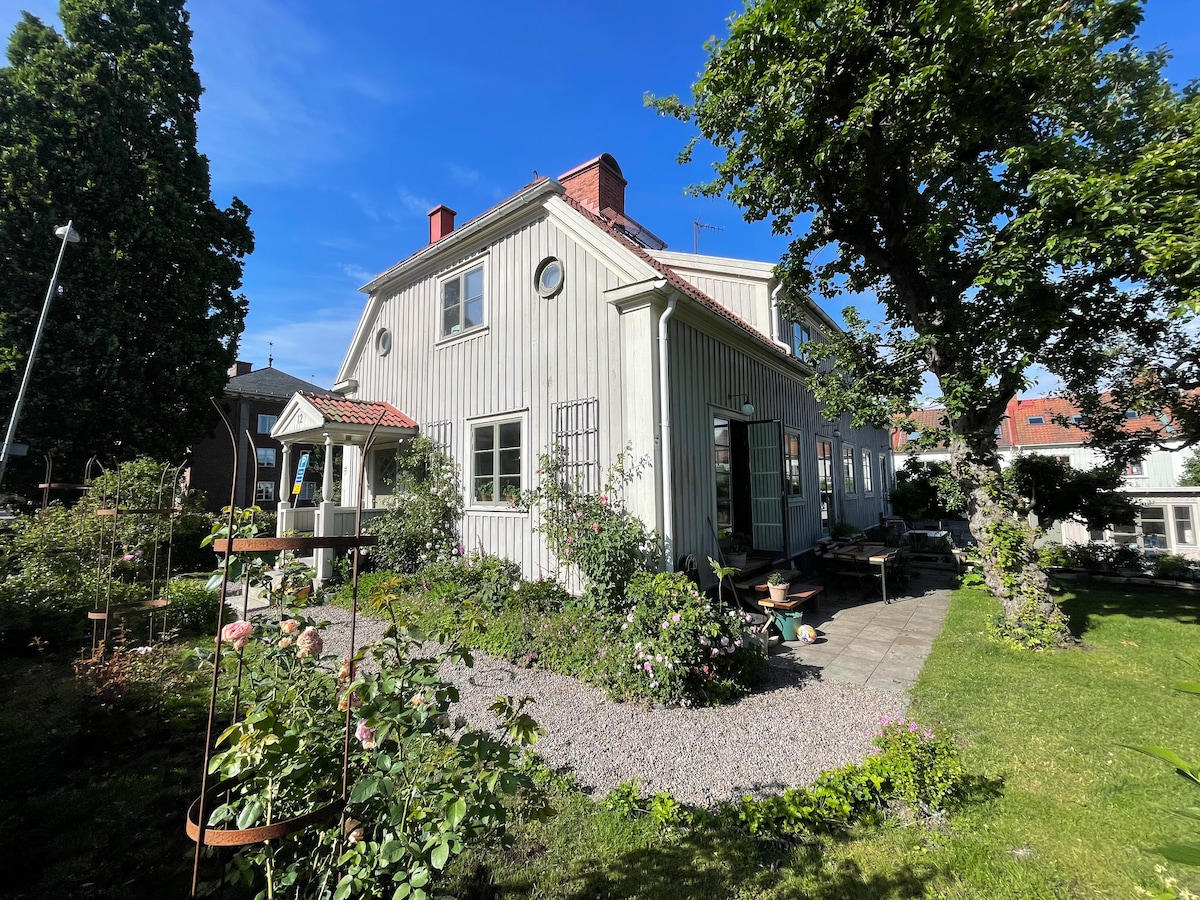 A two-story house with light-colored wooden siding is placed in a lush garden. Surrounding greenery includes trees and flowering plants. A stone patio is visible, offering seating areas, with pathways leading through the garden. The clear blue sky enhances the inviting exterior.