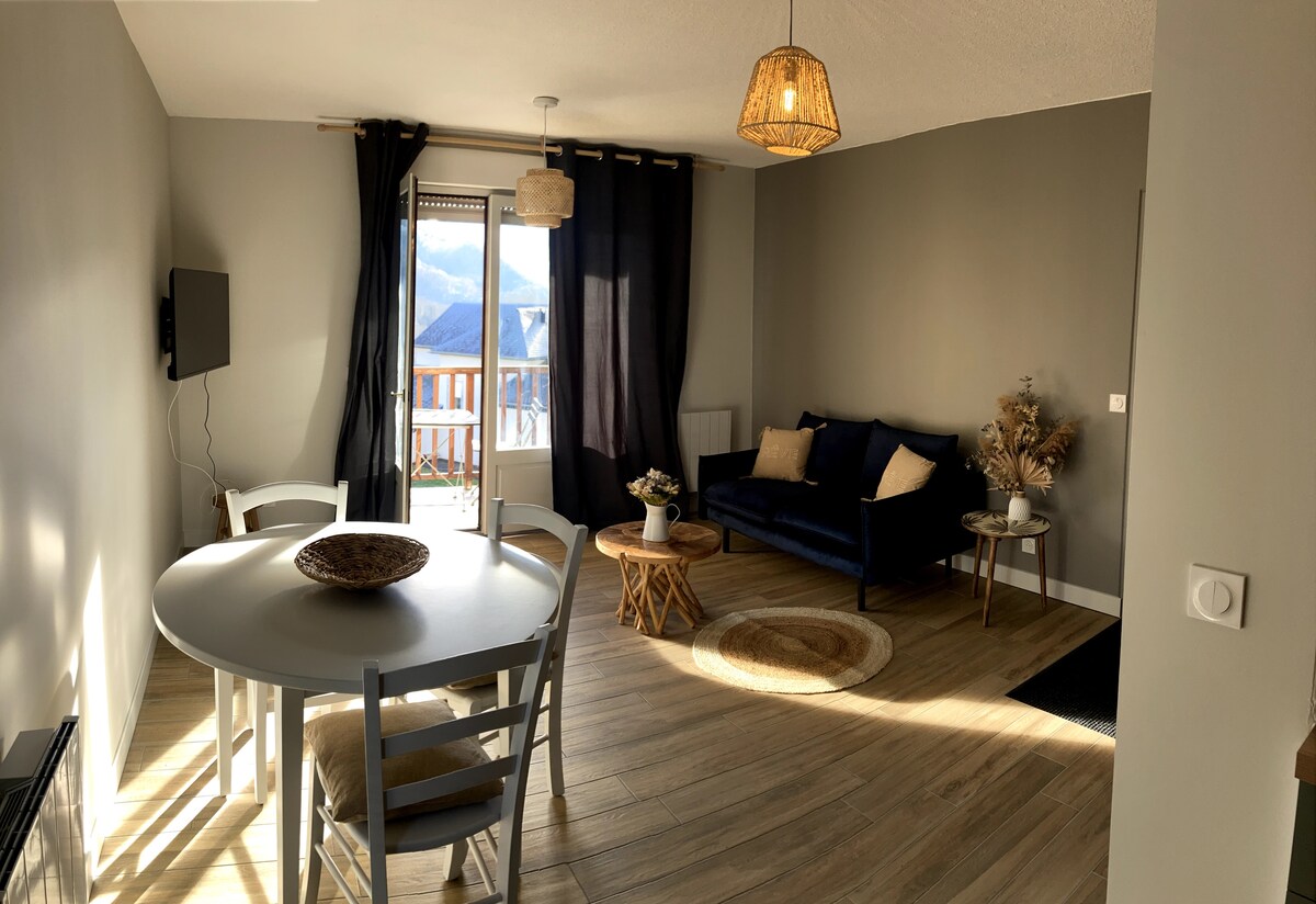 A light-filled living area features a round dining table surrounded by four chairs and a comfortable sofa with decorative cushions. A woven light fixture hangs from the ceiling, while a window with dark curtains offers scenic views of the outdoors.