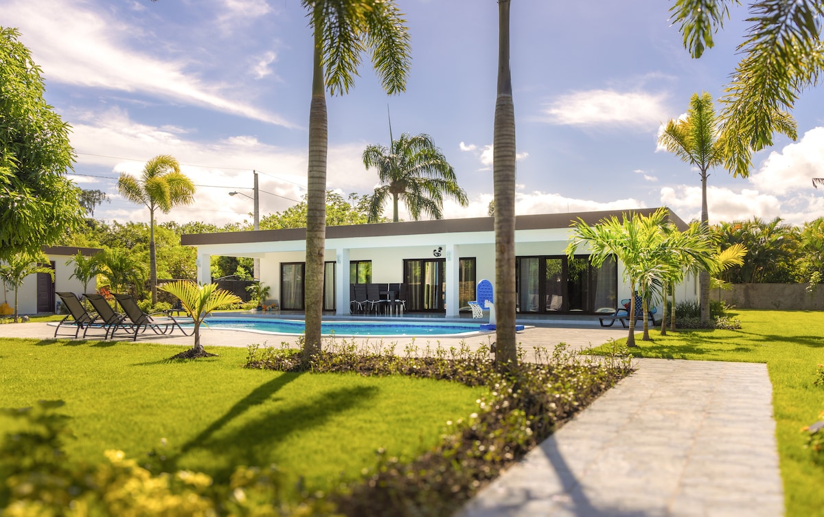 A spacious villa is surrounded by lush gardens and palm trees, showcasing a large pool at the center. Comfortable loungers line the poolside, while sliding glass doors provide views into the inviting interior. A stone pathway leads through the manicured lawn towards the villa entrance.