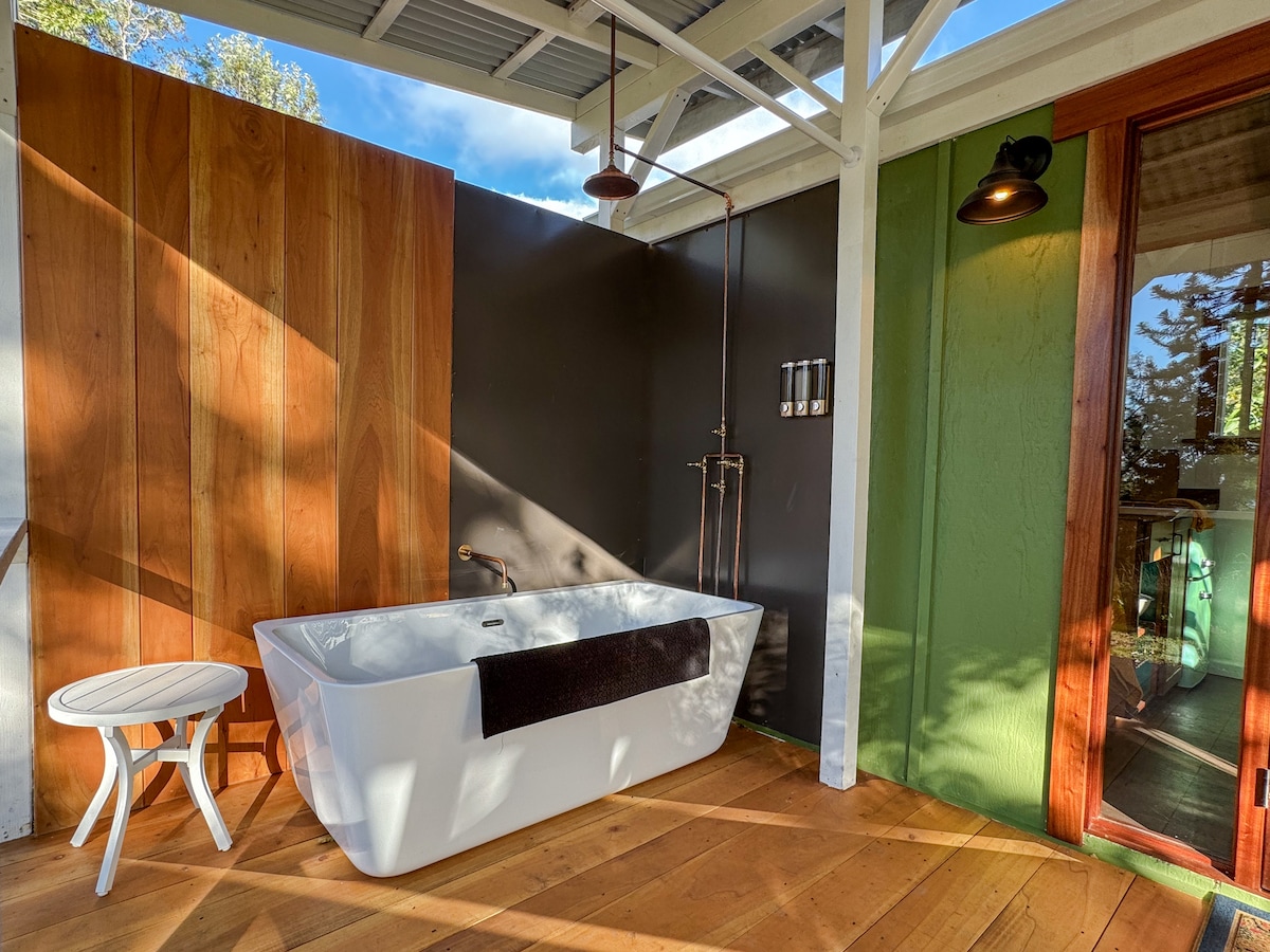 An outdoor soaking tub is positioned under a covered area, framed by wooden walls and surrounded by exposed decking. Natural light filters in, highlighting the clean lines of the freestanding tub and its sleek fixtures. A small white stool sits nearby, adding a functional touch to the serene space.