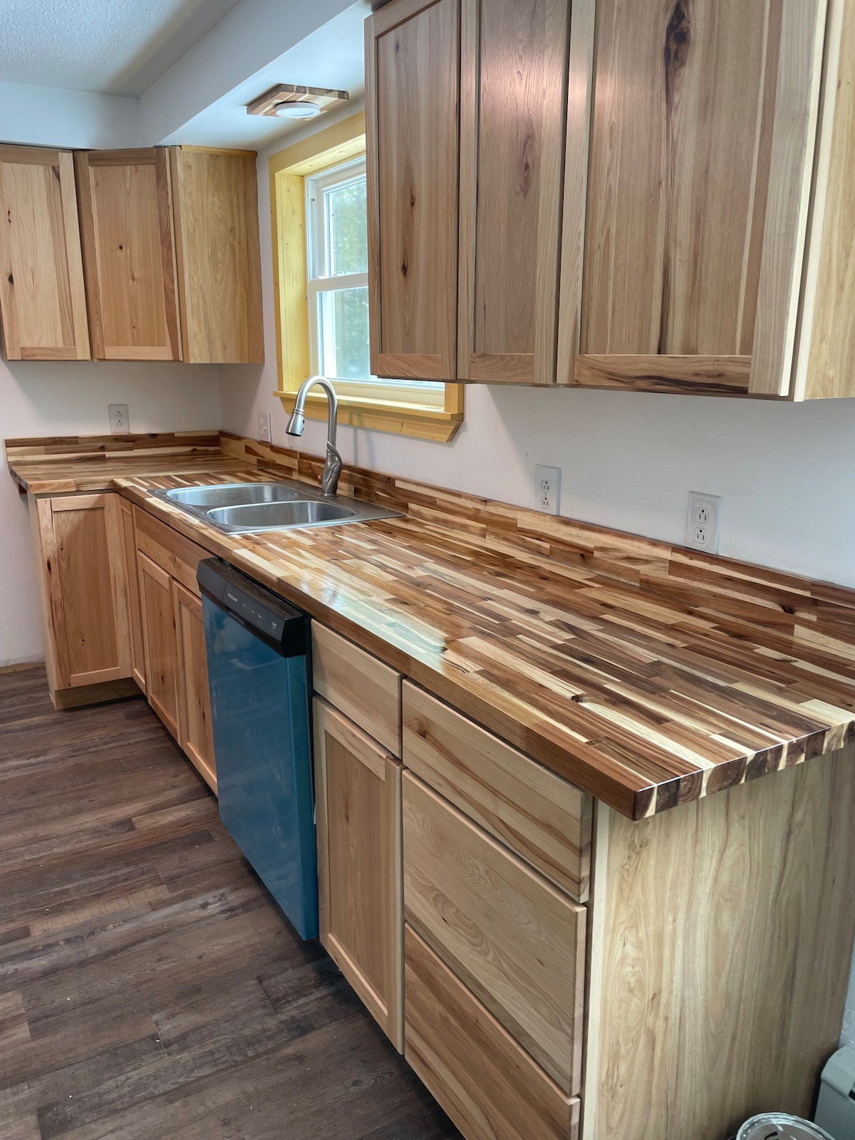 A modern kitchen showcases wooden cabinetry and a strikingly patterned countertop. A stainless steel sink is positioned under a window, allowing natural light to enhance the space. A blue dishwasher contrasts with the warm tones of the wood, creating a welcoming cooking area.