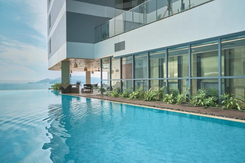 An inviting infinity pool is situated on the sixth floor, surrounded by lush greenery and modern lounge seating. Large glass windows reflect natural light, enhancing views of the sky and distant mountains. The tranquil water's edge is designed for relaxation in a scenic setting.