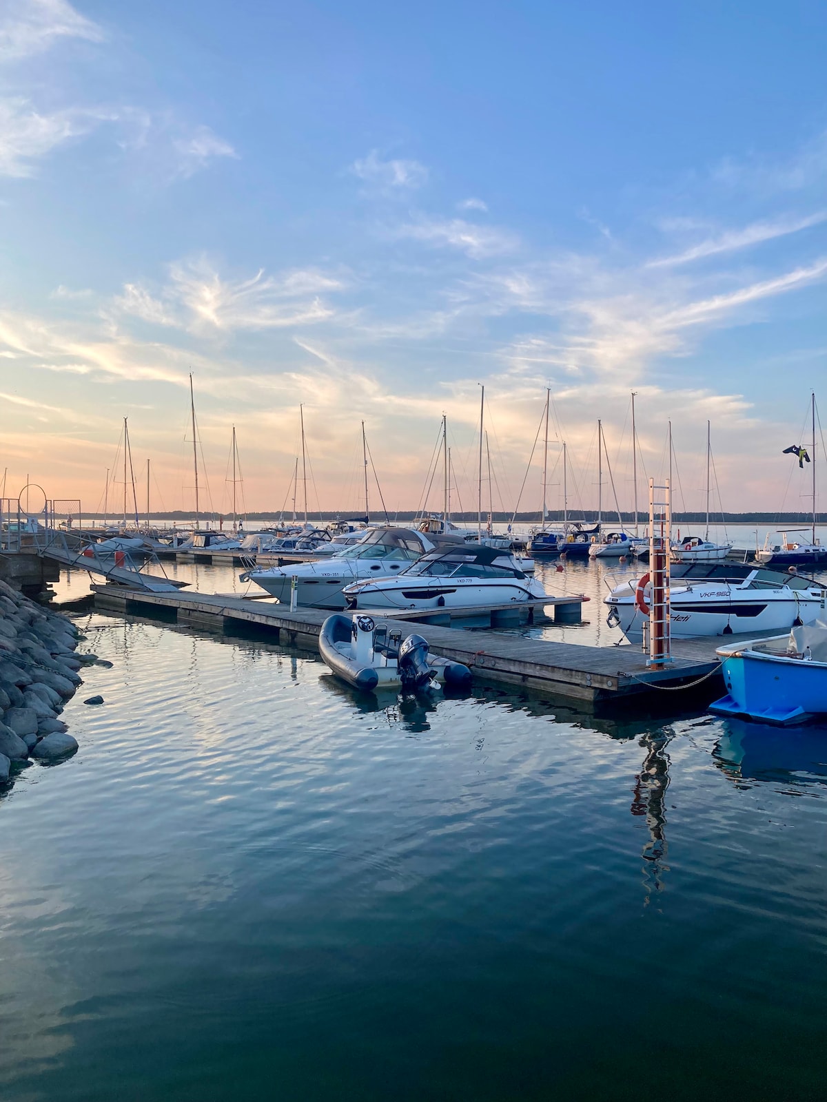 A serene marina scene is visible, showcasing various boats docked along a wooden pier. The calm water reflects soft hues of the sunset, while a modest ladder leans against the pier. Sailboat masts rise gently against a pastel sky.