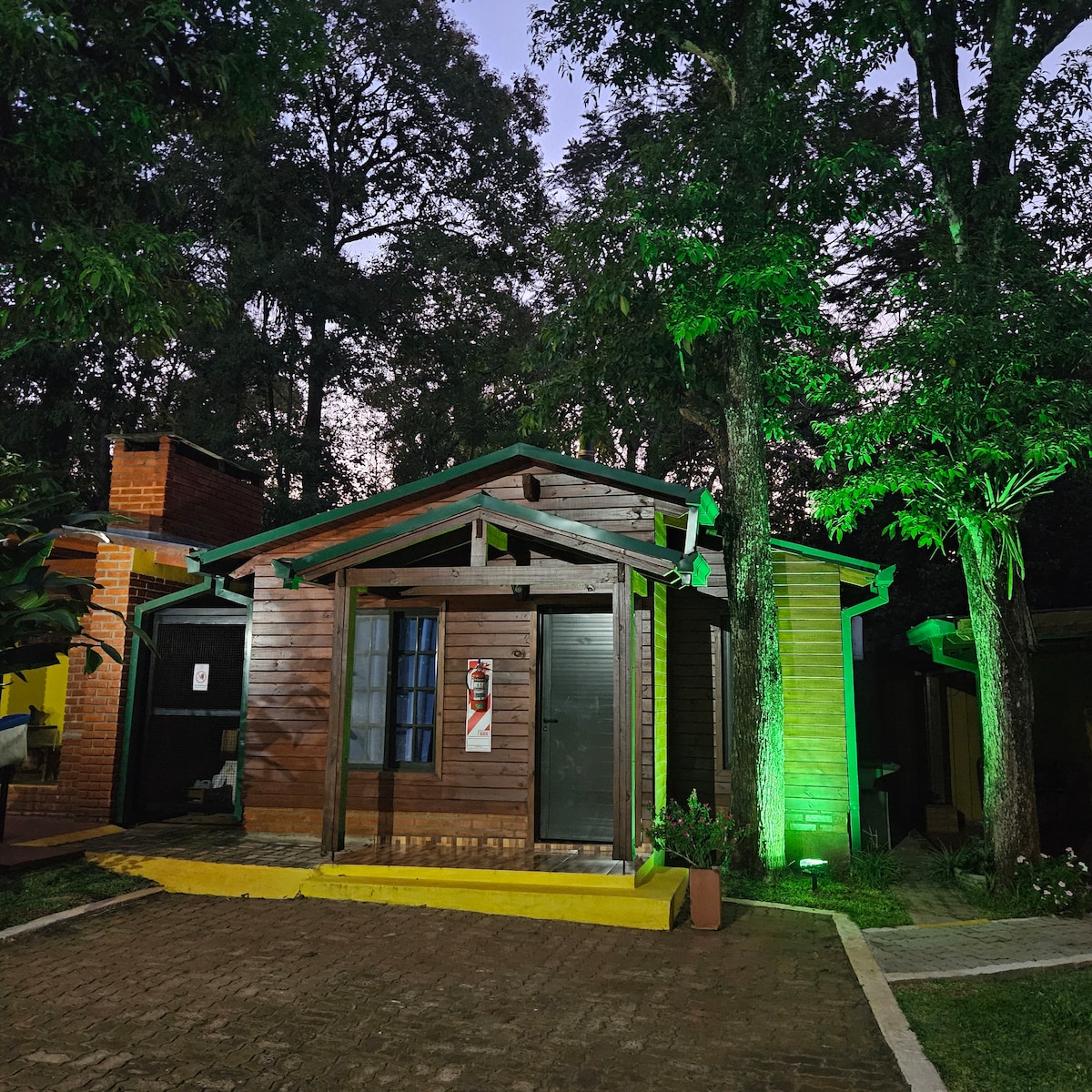 A wooden cabin is nestled among trees, illuminated by soft green lighting. The structure features a front porch with a single window and a decorative banner. Paved stones lead to the cabin, while surrounding trees enhance the natural setting.
