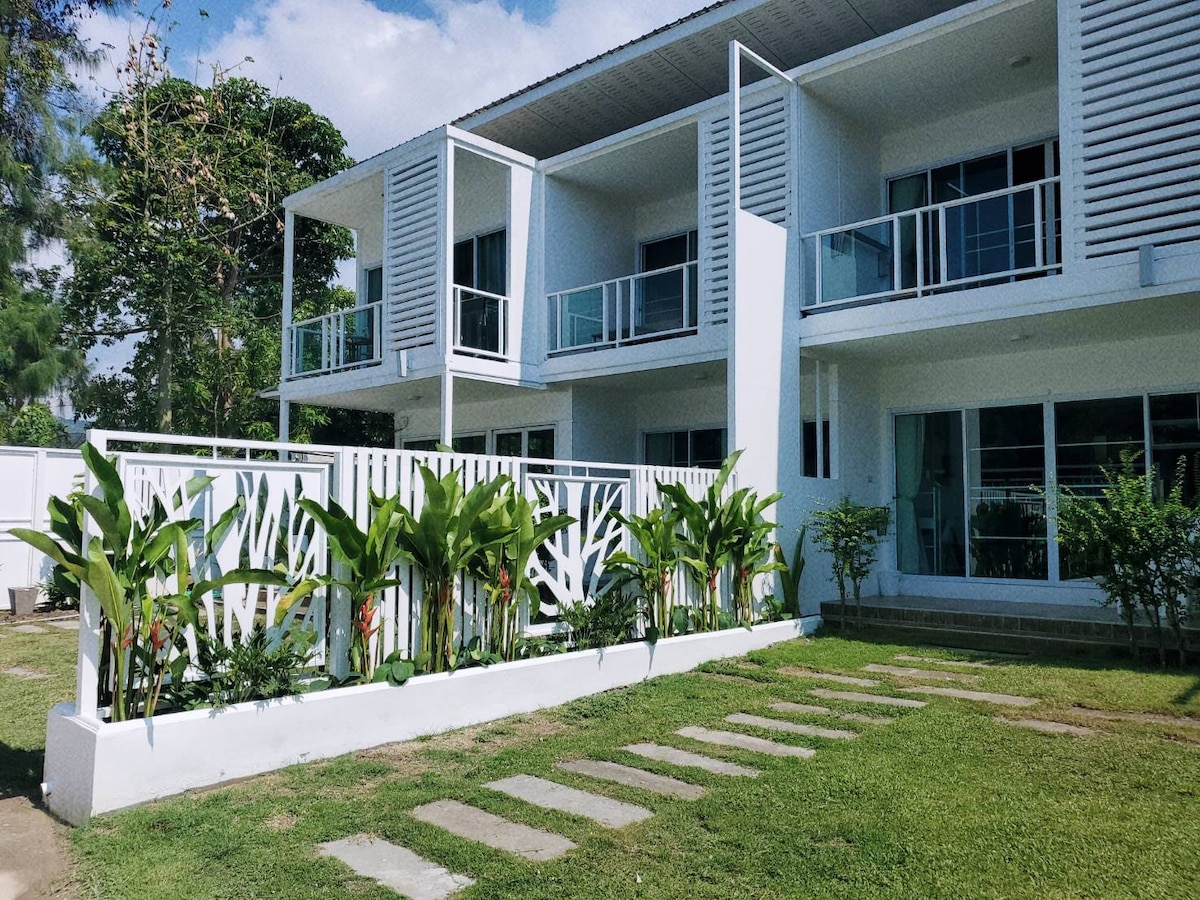 The exterior of the villa features a contemporary design with multiple balconies and large glass windows. Lush green plants frame the entryway, alongside a stone pathway leading to the front. The structure is set within a well-maintained garden area under a clear blue sky.