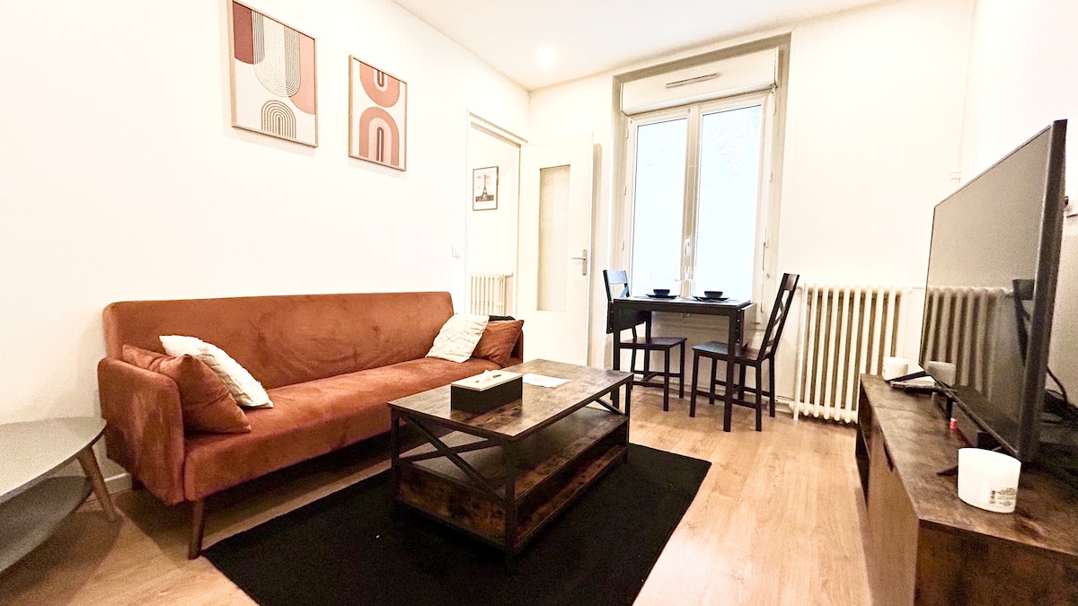 A living area is presented with a brown sofa adorned with decorative pillows. A wooden coffee table sits atop a dark rug, complemented by a flat-screen television. Natural light enters through large windows, illuminating a small dining table with chairs in the background.