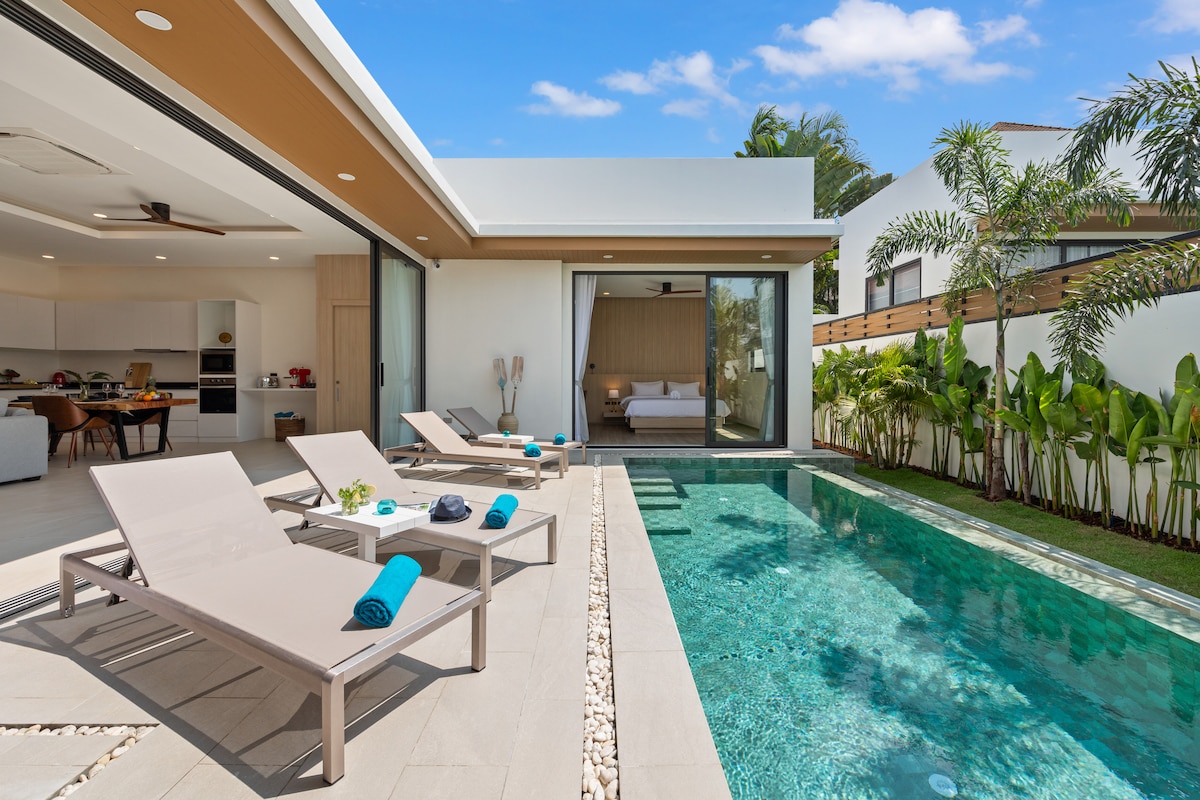 A serene outdoor space features a refreshing pool surrounded by light-colored loungers with turquoise towels. Sliding glass doors provide views into a spacious interior where natural light enhances the atmosphere. Lush tropical plants line the property, contributing to a private and inviting environment.