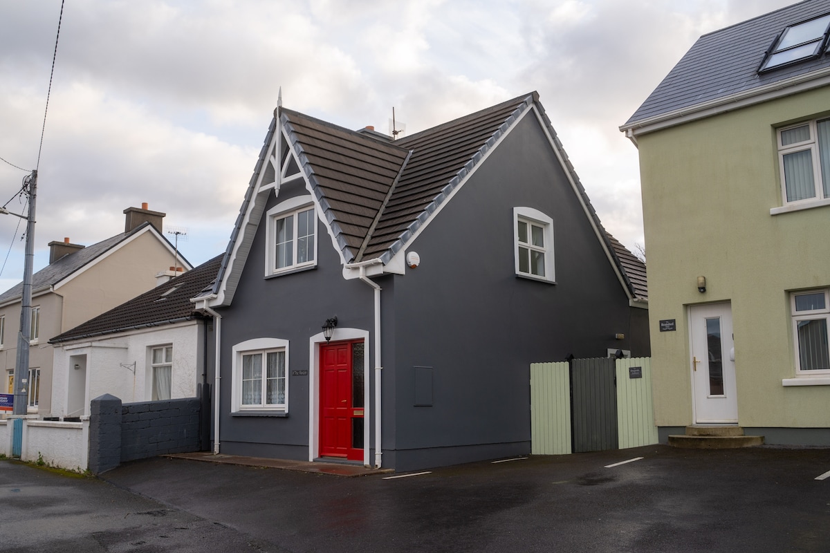 A charming two-story holiday home is shown, featuring a distinctive grey exterior with a red front door. The structure is flanked by neighboring homes and bordered by a low wall, with off-road parking visible directly in front.
