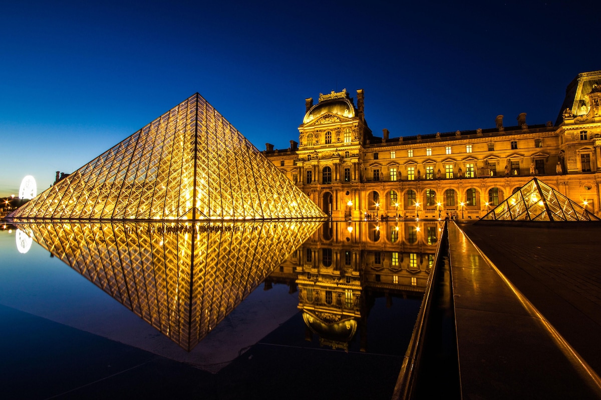 The Louvre Museum is illustrated with its iconic glass pyramid reflecting in the water at dusk. Illuminated lights cast a warm glow on the ornate architecture of the building, creating a striking contrast against the evening sky.