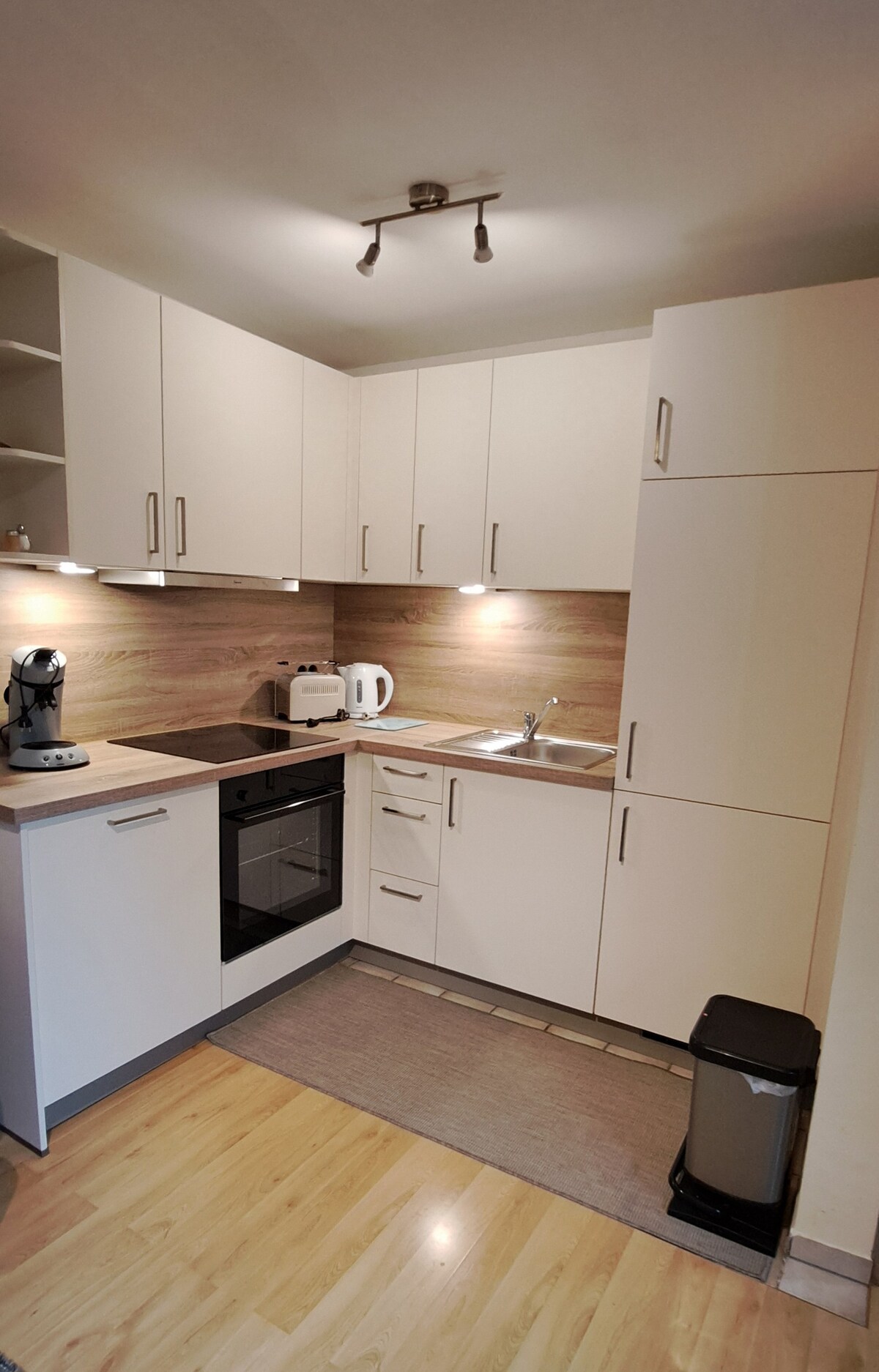 A modern kitchen features sleek cabinetry with a light wood finish, equipped with essential appliances including an oven, kettle, and coffee maker. Under-cabinet lighting enhances the workspace, while a small rug defines the cooking area on a light wooden floor.