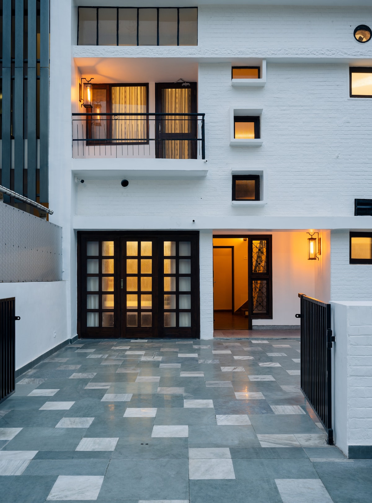 The welcoming entrance features a spacious, well-lit courtyard with patterned stone flooring. The contemporary facade is highlighted by large glass doors and strategically placed windows, inviting natural light. Warm lighting fixtures add a subtle glow, creating an inviting atmosphere for guests.