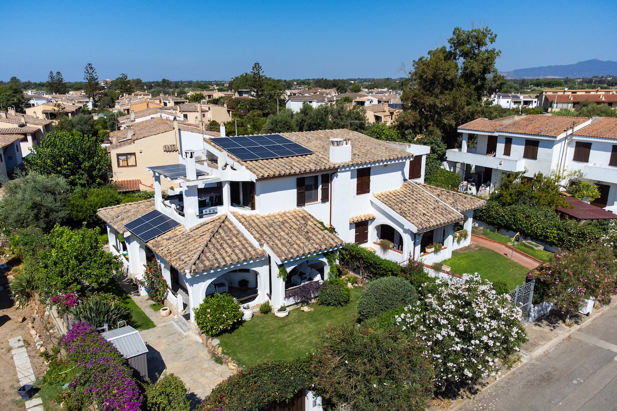 A spacious villa is showcased from above, featuring a terracotta-tiled roof and solar panels. The lush garden is surrounded by colorful flowers and greenery, while pathways lead to inviting outdoor seating areas, offering a peaceful retreat in a serene neighborhood setting.