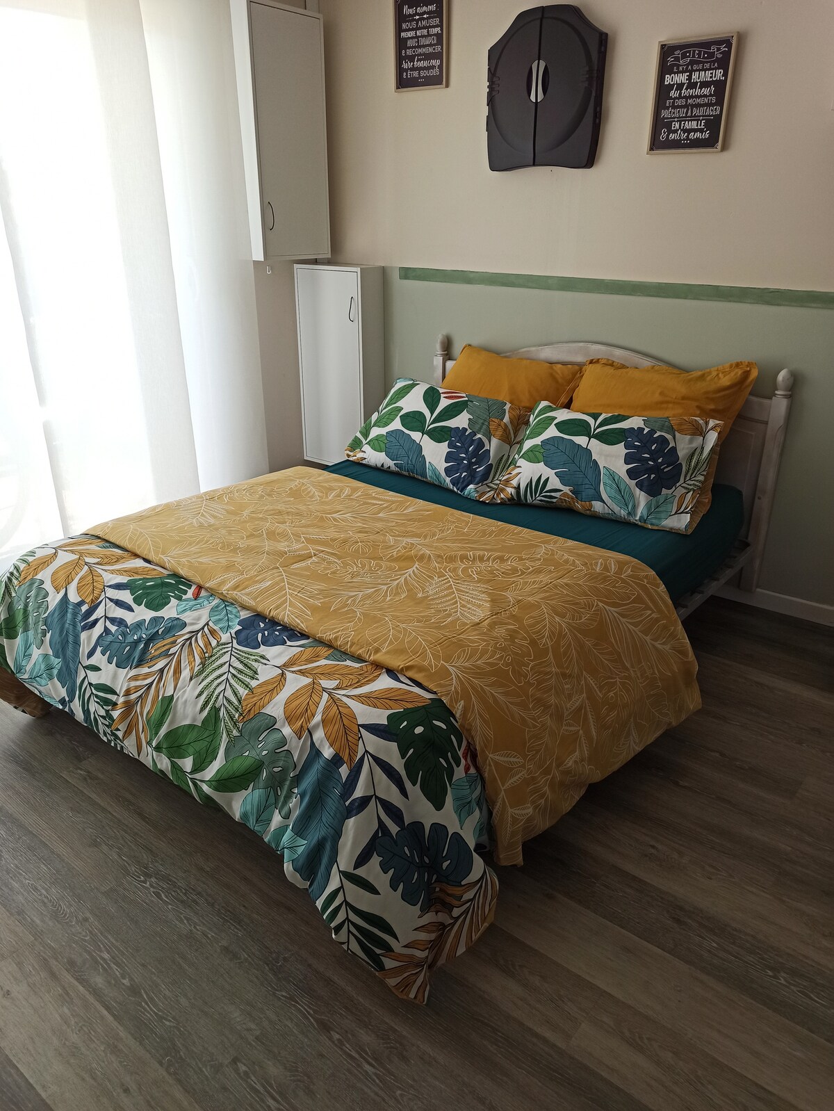 A comfortable sleeping area features a bed dressed in a bright, floral-patterned duvet and matching pillows. Natural light filters through sheer curtains, lending a soft glow to the room. Storage cabinets are placed conveniently along one wall.