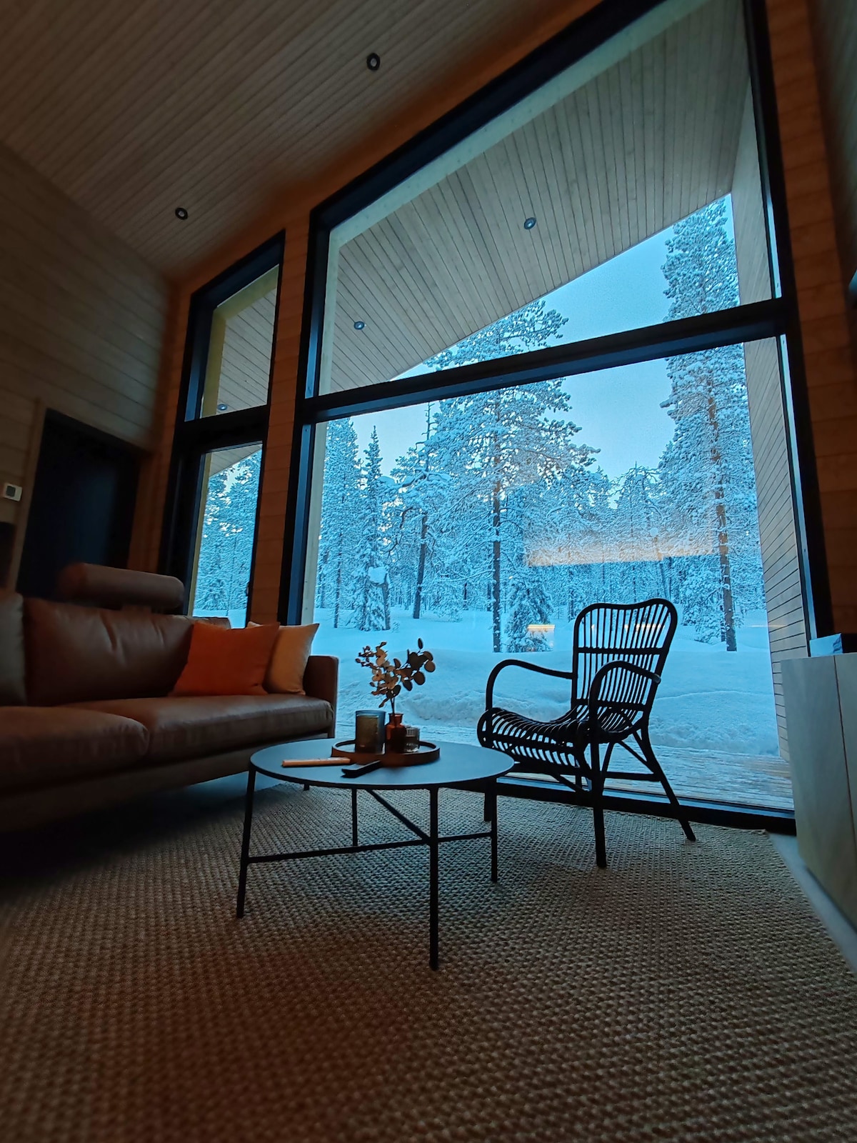 A spacious living area features large glass windows allowing views of a snowy landscape filled with tall trees. A sleek sofa is positioned next to a small coffee table, while a black chair offers additional seating, creating a cozy atmosphere.