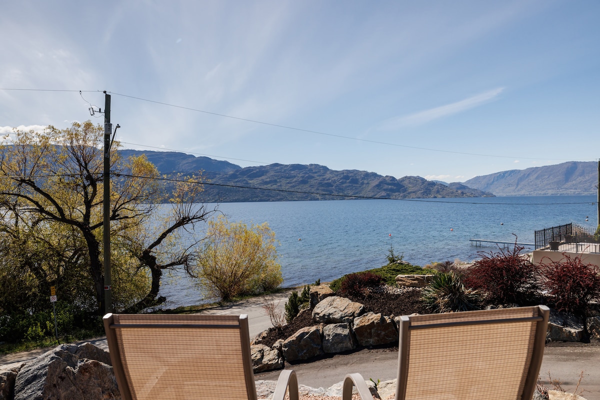 Lakeside views are captured from a seating area featuring two lounge chairs. The serene Okanagan Lake is visible, framed by gentle mountains in the background. A clear blue sky and soft clouds complement the peaceful atmosphere, while greenery and landscaped areas add to the natural setting.