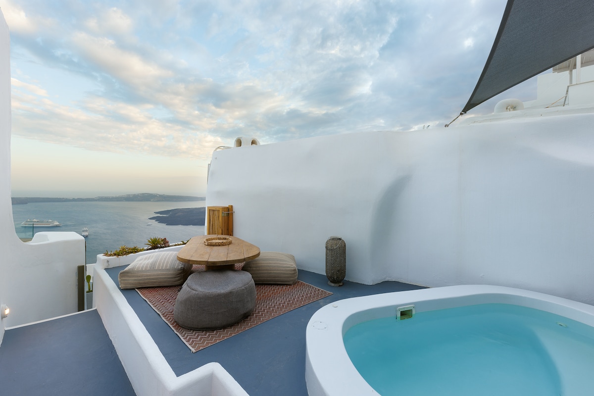 A serene outdoor space features a private Jacuzzi surrounded by smooth white walls. A low table with a round top is set upon an area rug, with large cushions for seating nearby. The distant horizon meets the tranquil sea under a softly clouded sky.