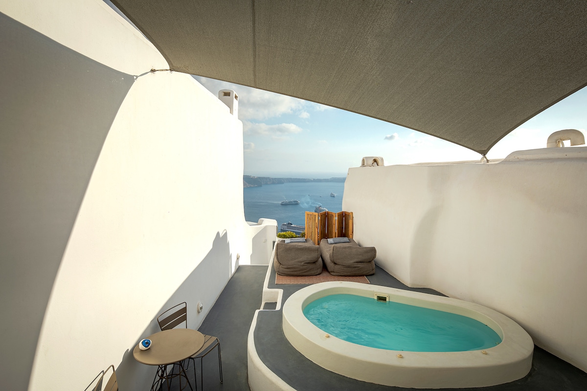 An outdoor area features a private jacuzzi surrounded by soft cushions and a low table, with expansive views of the sea and horizon. The space is shaded by a retractable awning while maintaining a modern aesthetic through the clean lines of the white walls.