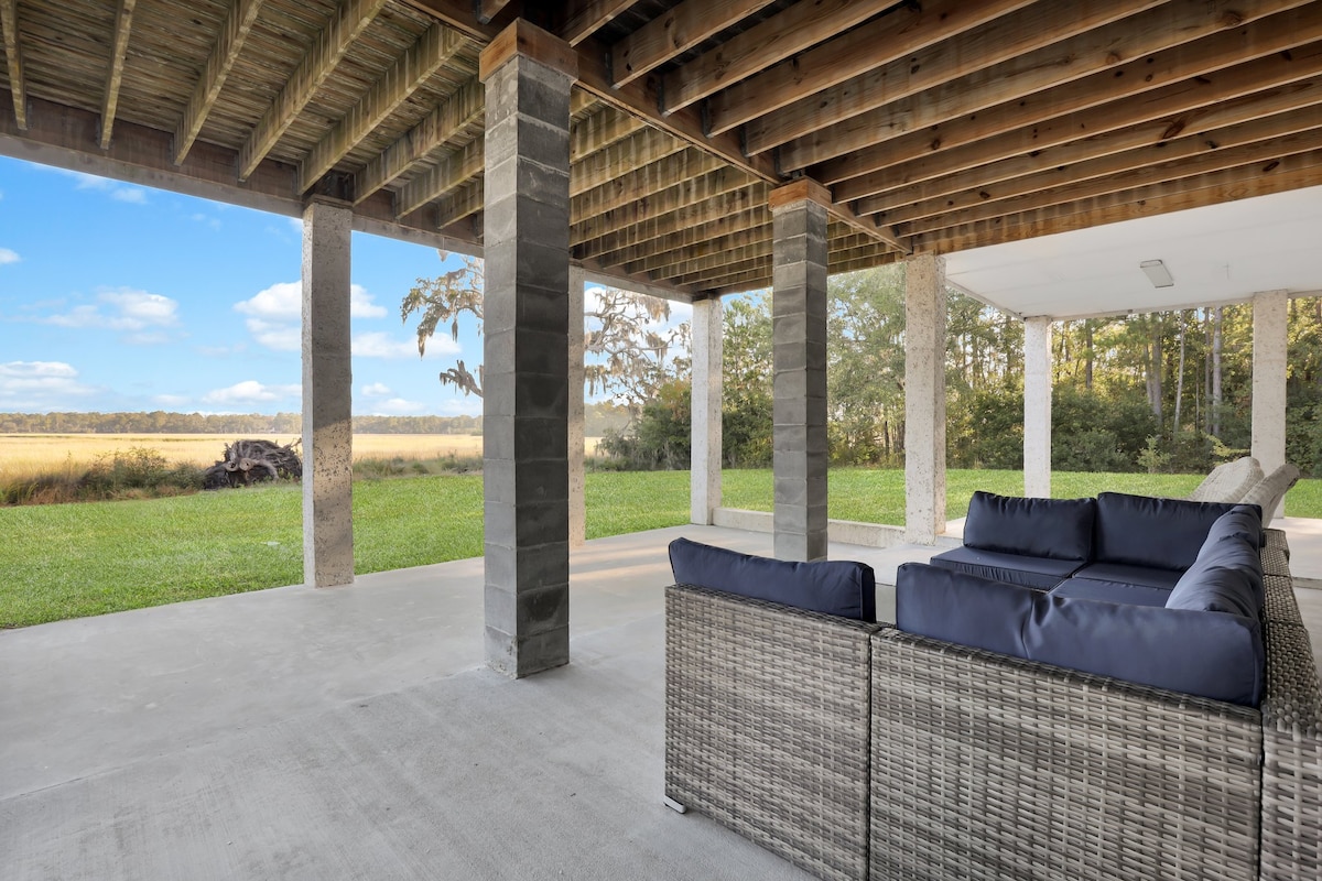 An outdoor seating area is presented under a covered patio, featuring a woven couch with dark cushions. The view extends to a lush green lawn and marshland, framed by trees and an open sky, creating a serene and spacious atmosphere.