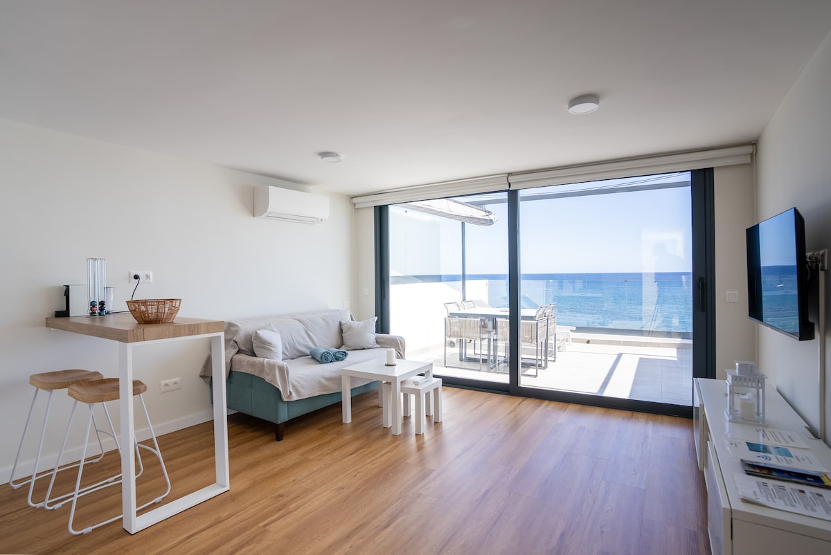 A bright living area is featured with large glass doors that open to a balcony overlooking the sea. A comfortable sofa is positioned next to a minimalist table and stylish chairs. Light wood flooring complements the clean, modern design of the space.