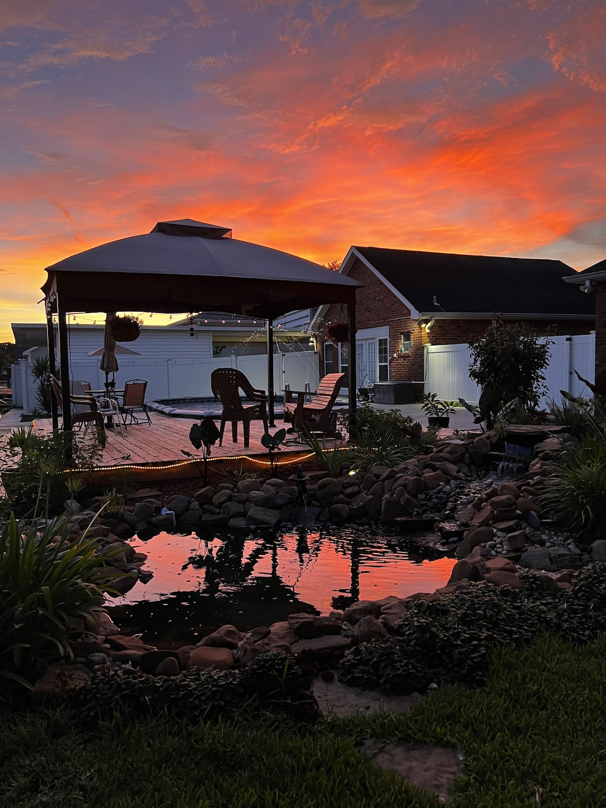 A serene backyard scene features a wooden deck with seating beneath a gazebo against a vibrant sunset. The tranquil pond reflects the colorful sky, surrounded by lush greenery and rocks, creating a peaceful outdoor setting.