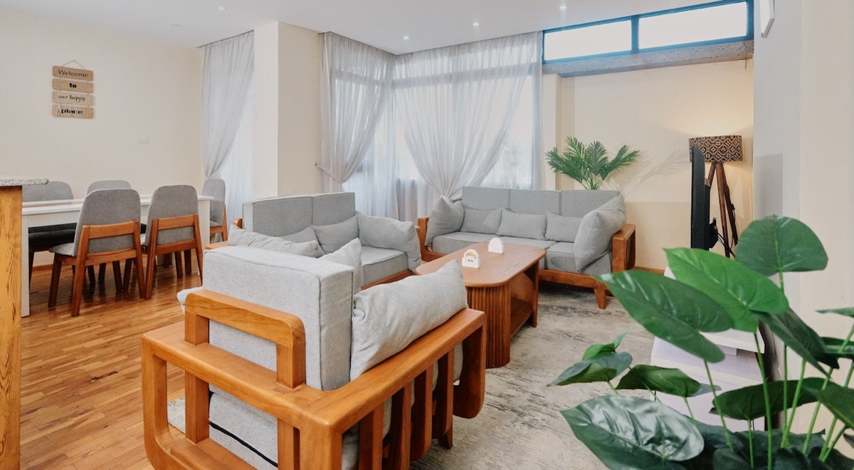 A spacious living area features soft grey couches arranged thoughtfully around a wooden coffee table. Natural light floods in through large windows, with sheer curtains gently diffusing the brightness. A decorative plant in the foreground adds a touch of greenery to the modern decor.