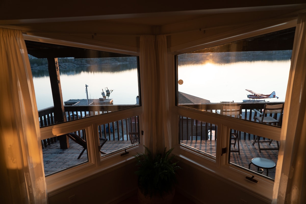 Panoramic windows frame a tranquil view of the water, with a float plane visible at the dock. The warm light of the sunset casts a soft glow on the deck furniture outside, suggesting a serene atmosphere for relaxation.