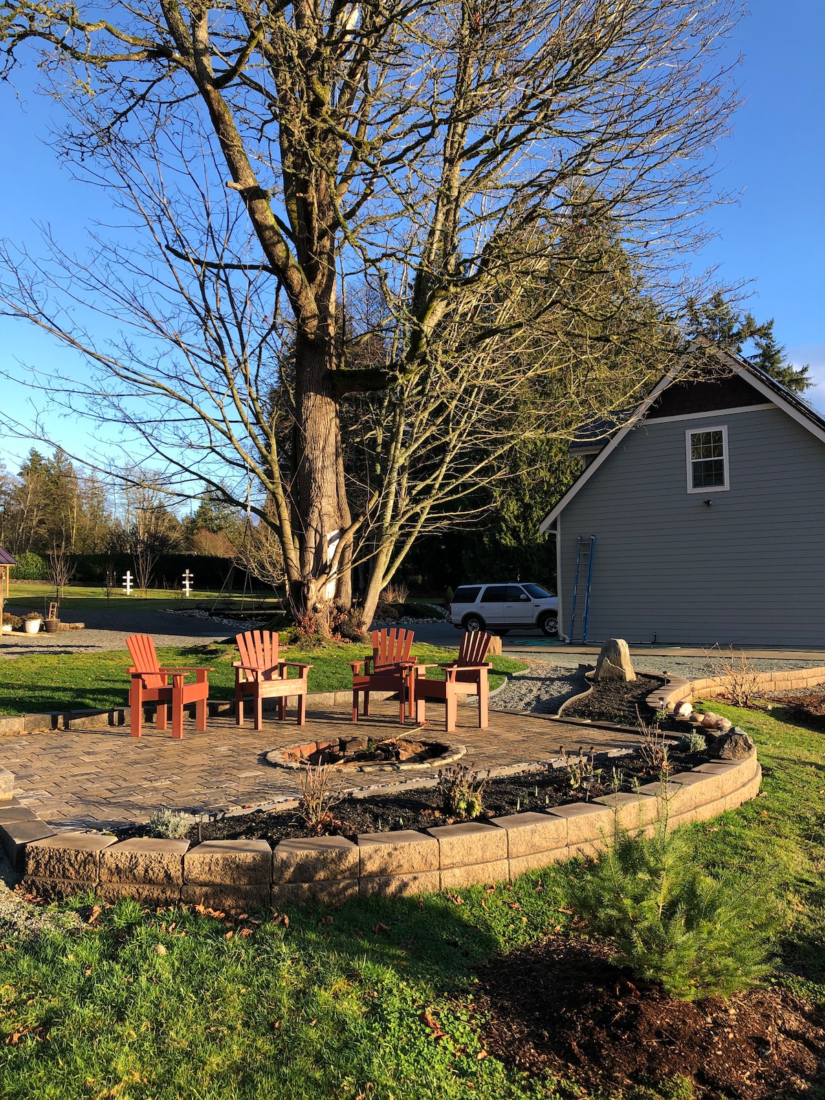 A circular stone patio features four Adirondack chairs arranged around a fire pit. A large tree provides shade, while a lawn area and garden beds are visible nearby. A two-story building is partially shown in the background with a vehicle parked close by.