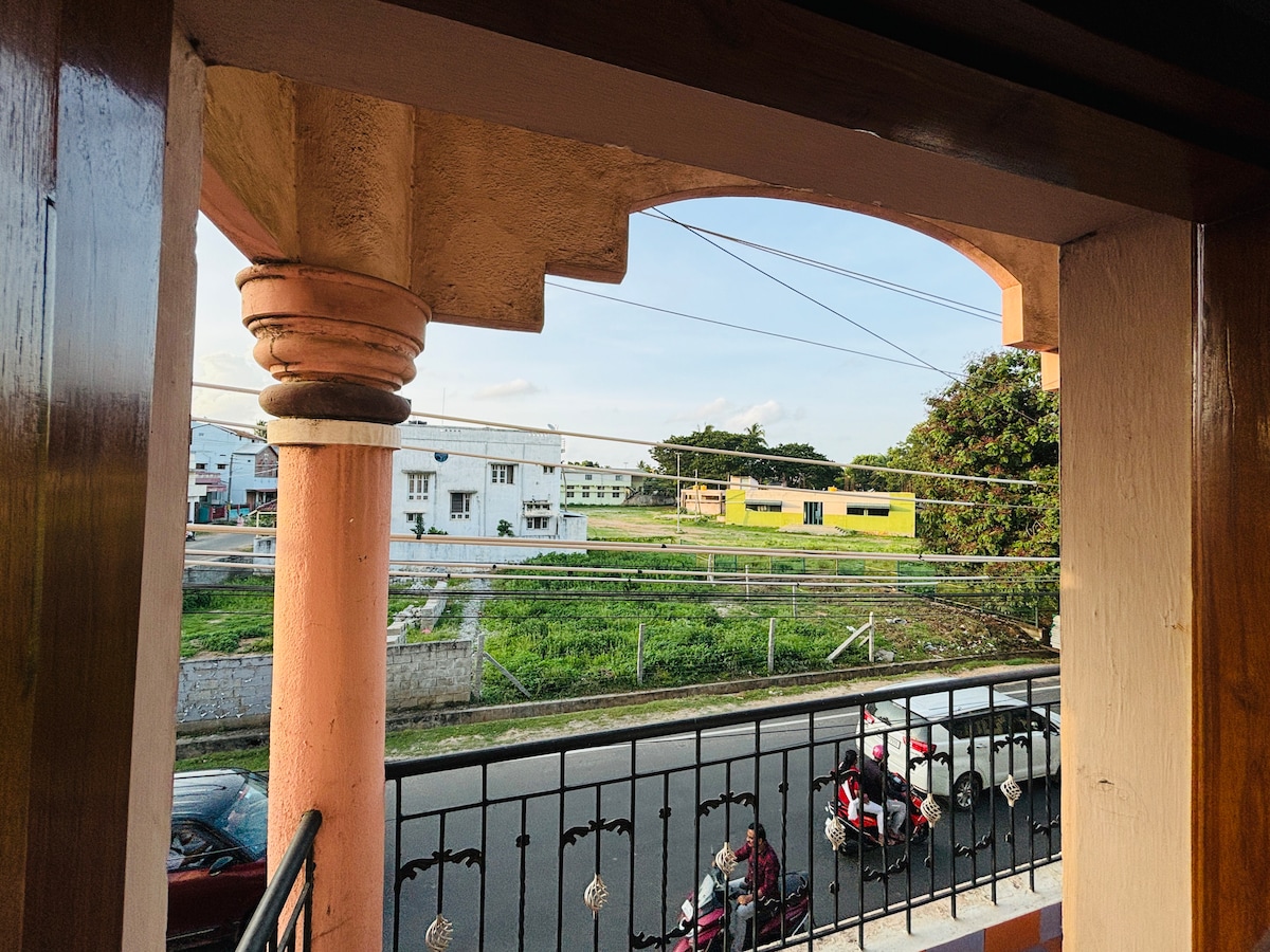A view is captured from a balcony featuring decorative pillars. The scene includes a road lined with parked vehicles and a grassy area across the street, complemented by structures in the background under a clear sky.