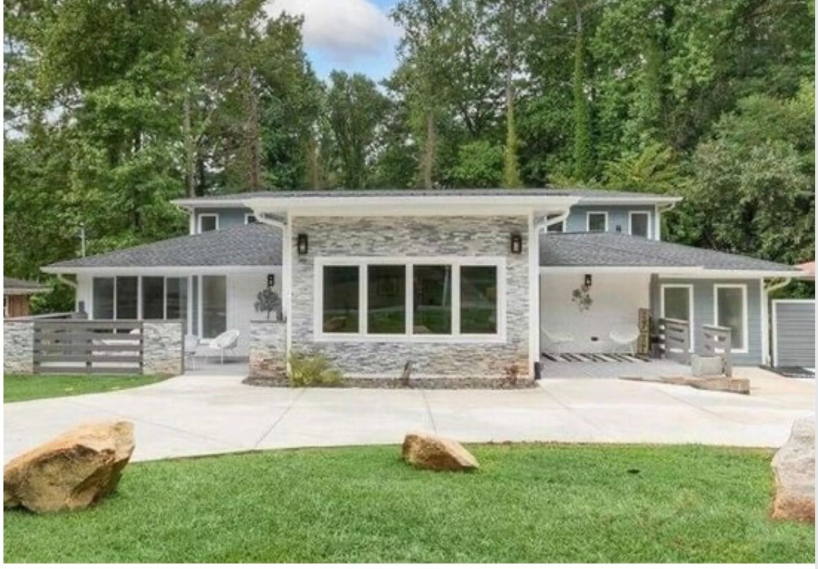 A spacious exterior view of the studio highlights a modern design with large front windows, allowing natural light to brighten the interior. Green grass surrounds the home, while natural stone accents and a paved pathway create an inviting entrance.