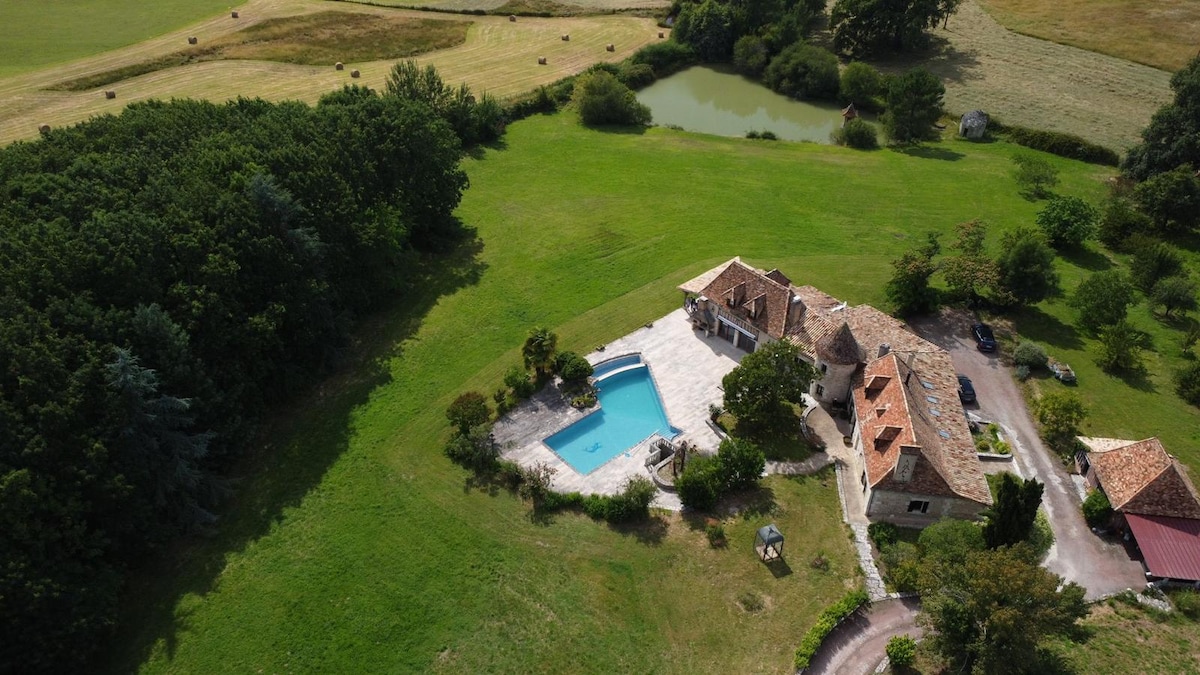An aerial view showcases a spacious rural property surrounded by lush greenery and open fields. A rectangular swimming pool is visible adjacent to a terrace area, while the house features rustic architecture with a tiled roof. A tranquil pond is seen nearby, enhancing the natural setting.