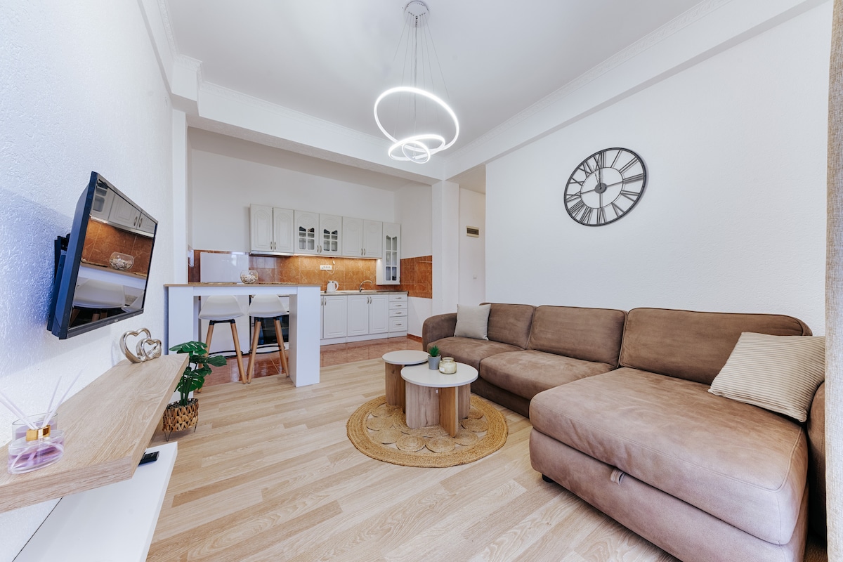 A spacious living area features a large brown sectional sofa arranged around a circular coffee table. A modern light fixture hangs overhead. The kitchen area, visible in the background, is equipped with white cabinetry and wood accents. A wall clock adds a decorative touch.