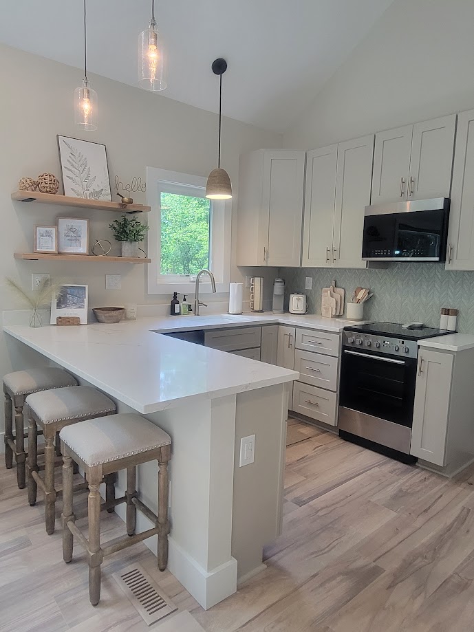 The kitchen features sleek cabinetry and modern stainless steel appliances. A well-lit island with three upholstered stools offers seating, while natural light illuminates the space through a large window. Decorative elements and utensils are neatly arranged on open shelves, creating a functional and inviting atmosphere.