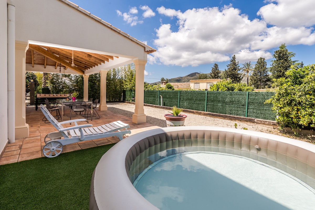 An outdoor area features a round hot tub, surrounded by grass and patio tiles. A shaded porch holds a dining table and chairs, offering space for relaxation. Lush greenery and a privacy fence are present in the background, with distant hills visible under a partly cloudy sky.