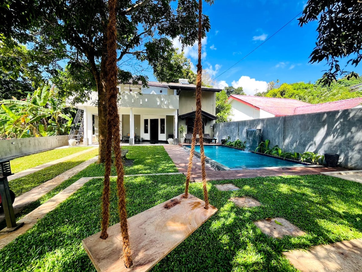 A serene outdoor space features a spacious lawn bordered by lush greenery. A wooden swing hangs from a sturdy tree, overlooking a clear swimming pool. The two-story building with a light-colored exterior is visible in the background, complemented by a bright blue sky.