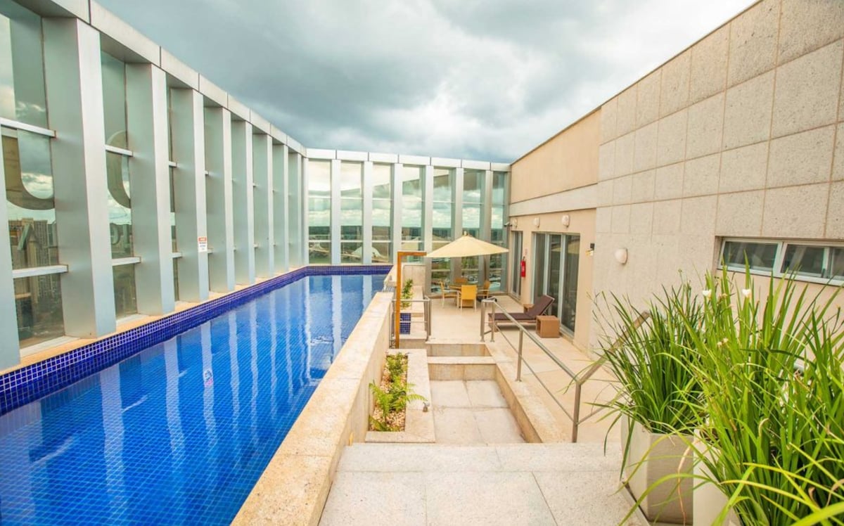 A rooftop pool is seen surrounded by lounge chairs and an umbrella for shade. Large glass panels provide views of the city, while a well-maintained garden area is visible nearby. The tranquil atmosphere invites relaxation and leisure.
