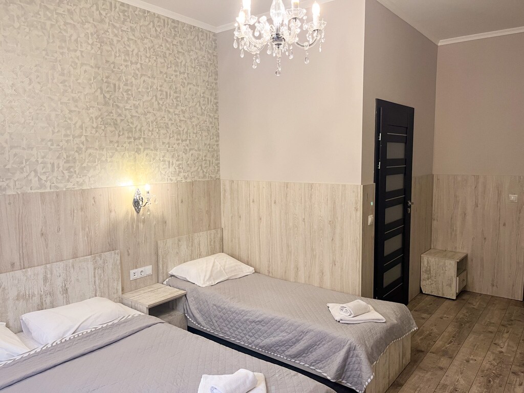 A guest room features two spacious beds, each adorned with light grey bedding. Wall sconces provide soft lighting alongside a decorative chandelier. Light wood paneling accents the walls, with a simple storage bench located near the door.