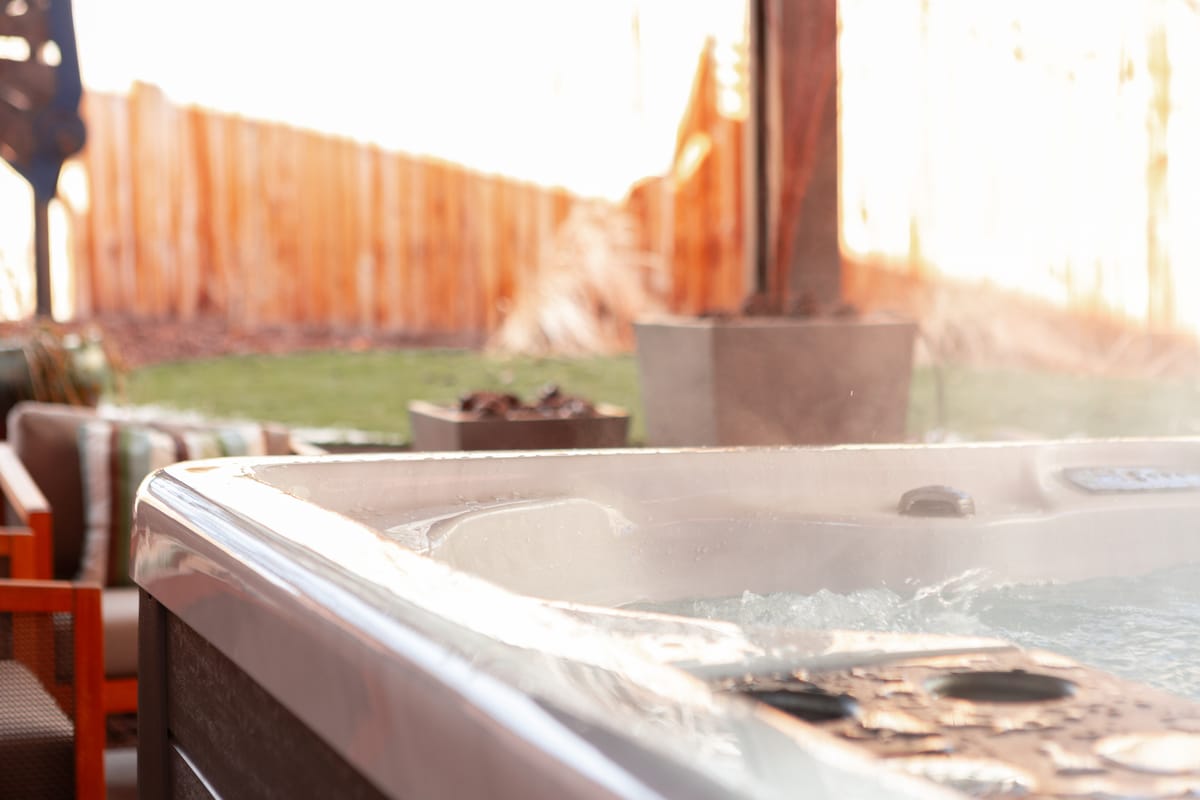 A new hot tub is shown at the edge of a patio, with steam rising gently from the water's surface. Comfortable seating is visible in the background, accentuated by a lush green lawn and wooden fencing that provides privacy.
