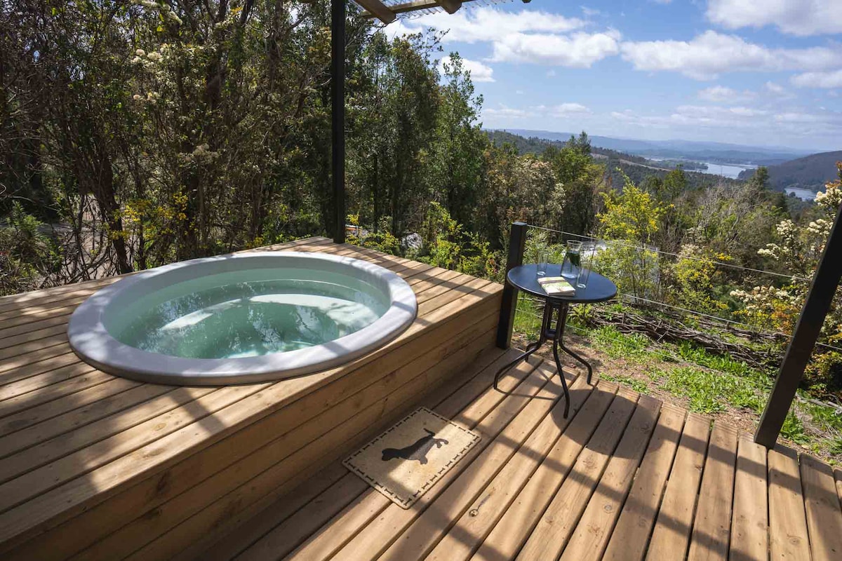 A private hot tub is positioned on a wooden deck, offering views of the surrounding forest and rivers. A small table set for two, with wine glasses, complements the outdoor space, inviting relaxation amidst nature.